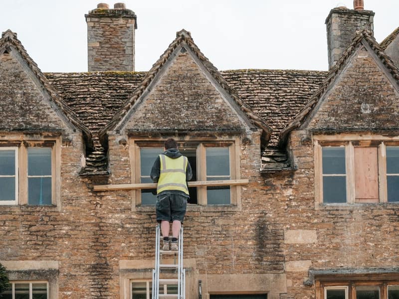 A roofer on a ladder inspecting the roofline of an older stone terrace during a pre-purchase roof check