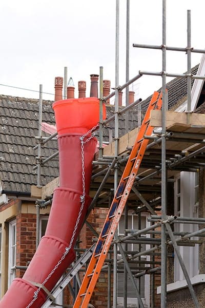 A red enclosed rubbish chute running from scaffolding down to ground level outside a brick terraced house