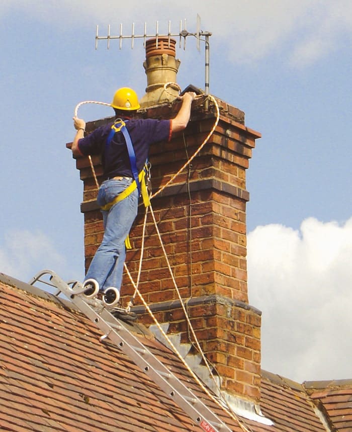 A roofer in a hard hat and harness working from a roof ladder hooked over the ridge of a tiled roof beside a brick chimney